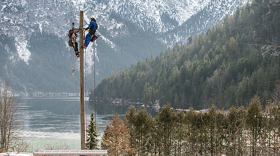 Techniker arbeitet auf Strommast in alpiner Landschaft nahe See